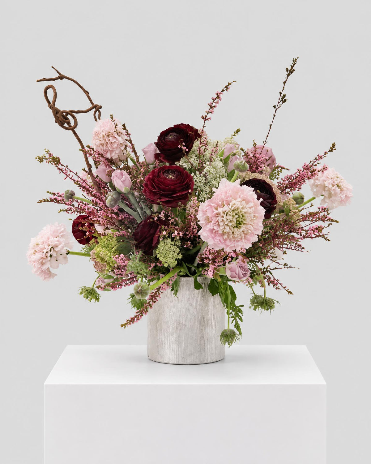 Blush Nocturne arrangement with burgundy ranunculus, blush pink blooms and twisting branches in a textured pale vase.